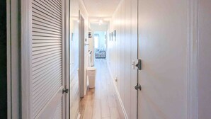 Un couloir étroit aux murs blancs et au plancher en bois, qui mène à une salle de bain équipée de toilettes et d’un espace douche.