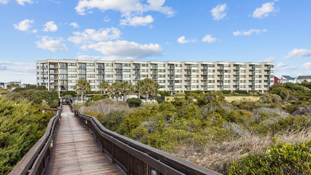 A wooden boardwalk leading to a multi-story building with balconies, surrounded by dense shrubbery.