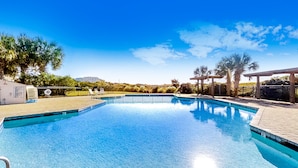 A clear blue swimming pool with a tiled deck, surrounded by palm trees and a covered seating area.