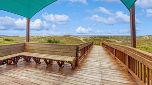 A wooden boardwalk with benches and a shaded area, leading to a beach with dunes and the ocean in the background.