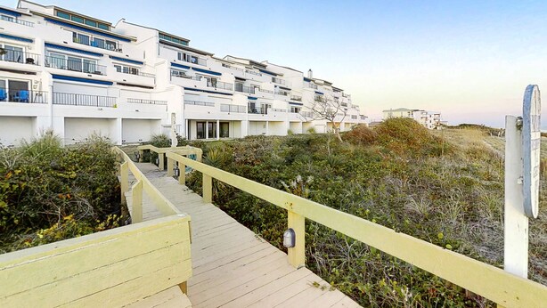 A modern multi-story building with balconies, a wooden walkway, and surrounding vegetation. - Oceanfront, 2nd-Floor Condo with Great Ocean View, Balcony, WiFi & Shared Pool (Pine Knoll Shores)