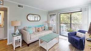 A living room with a beige sofa, a blue armchair, a wooden coffee table, and a large window with a view of greenery. - Condo by the ocean with a pool, tennis, WiFi, central AC - steps from the beach (Atlantic Beach)