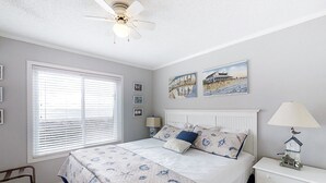 Ein Schlafzimmer mit Bett, Nachttisch, Lampe, Deckenventilator, Fenster mit Jalousien und Wandbildern.