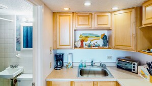 A kitchen with wooden cabinets, a sink, a coffee maker, a toaster, and a window with a decorative painting. - Shorely Suite #204: Waterfront suite, pool, beach access (Atlantic Beach)
