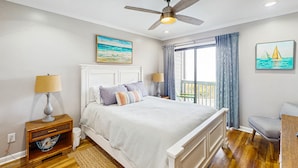 A bedroom with a bed, a ceiling fan, a nightstand with a lamp, a window with curtains, and a painting on the wall. - 2nd-Floor Condo in Oceanfront Location w/Shared Pool, Balcony, Free WiFi (Atlantic Beach)