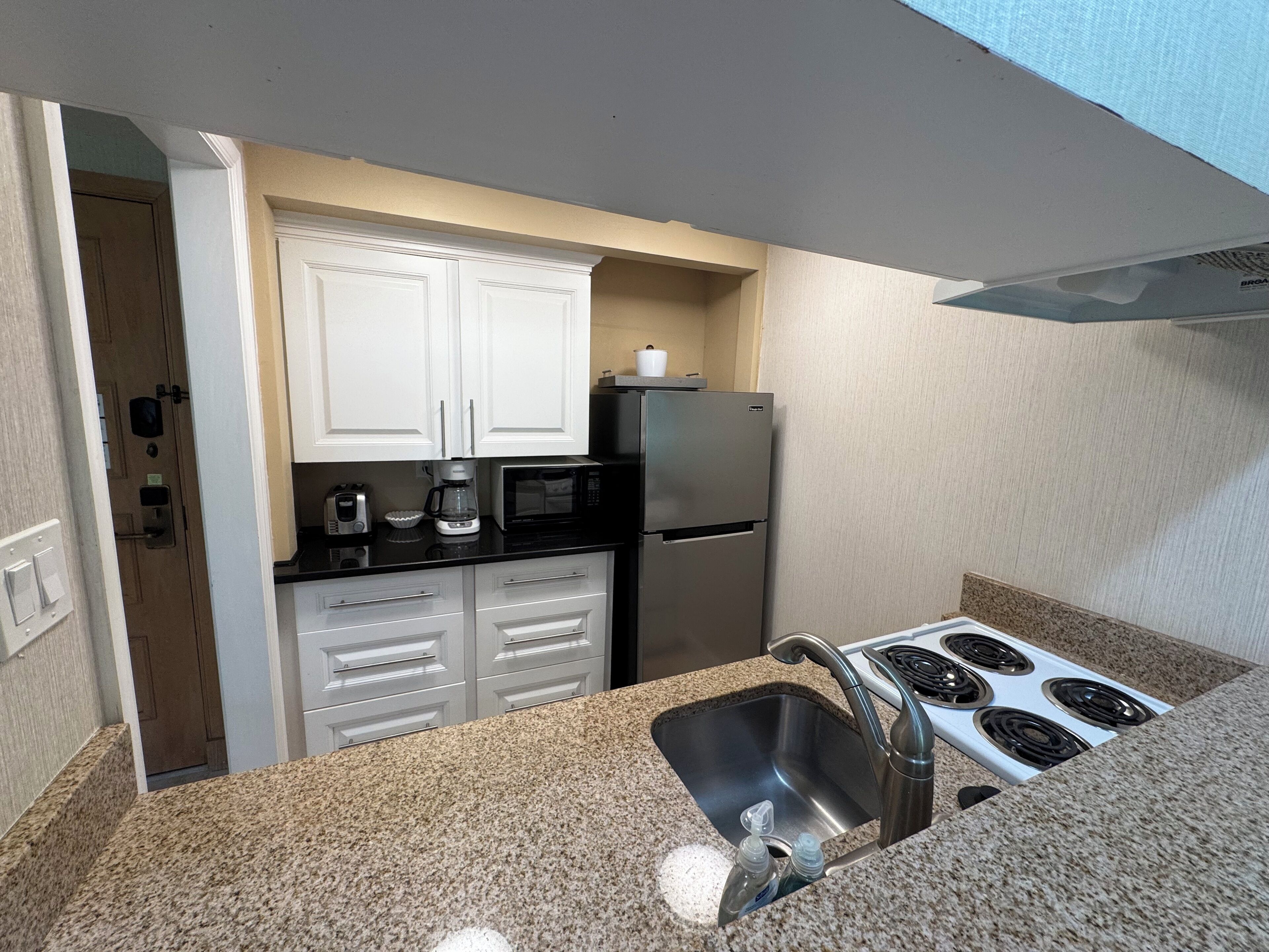 Cocina compacta con armarios blancos, pileta de acero inoxidable, microondas, refrigerador y estufa a gas.