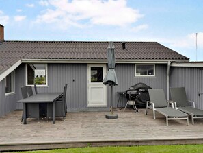 A grey metal building with a tiled roof, a covered patio area with a table and chairs, and a barbecue grill.