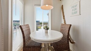 Une pièce avec une table blanche, des chaises en osier, un luminaire suspendu, une pancarte sur laquelle on peut lire « PRIEZ POUR QUE LES VAGUES SOIENT BONNES » et une vue par une fenêtre avec des stores.
