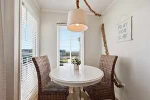 Une pièce avec une table blanche, des chaises en osier, un luminaire suspendu, une pancarte sur laquelle on peut lire « PRIEZ POUR QUE LES VAGUES SOIENT BONNES » et une vue par une fenêtre avec des stores.