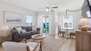 Un salon avec un canapé, des fauteuils, un téléviseur, un ventilateur de plafond et une vue sur l’extérieur, visible à travers une porte.