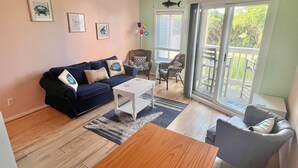 A living room with a dark blue sofa, a white coffee table, and a grey armchair. There is a large sliding glass door leading to a balcony with outdoor furniture. - First-Floor, Oceanfront Condo with WiFi & Shared Pools/Hot Tub/Tennis/Game Room (Atlantic Beach)