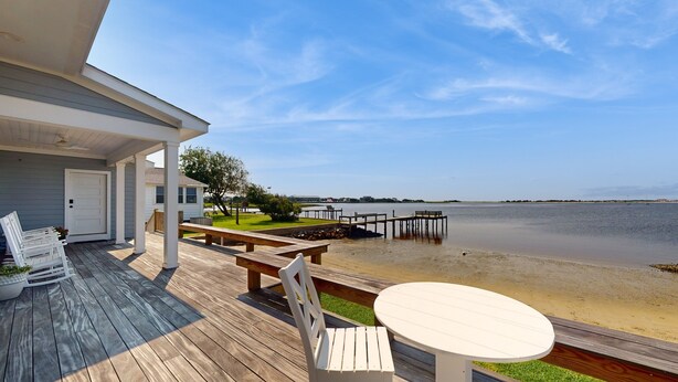 Une terrasse en bois avec des sièges d’extérieur, une table blanche et une vue sur un lac.