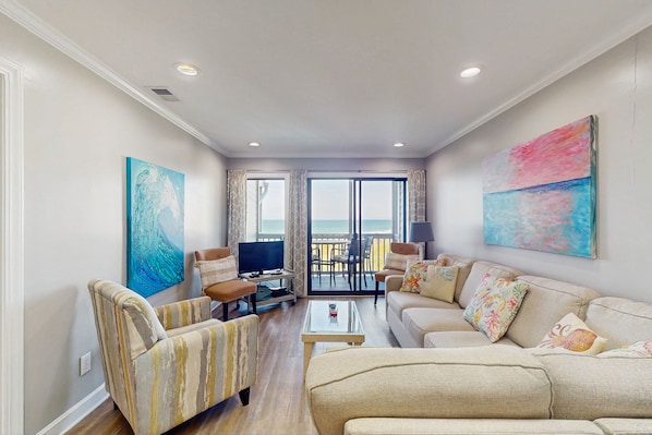 A spacious living room with a sofa, armchairs, a coffee table, and a television. There are large windows with curtains, a balcony, and artwork on the walls. - Third Floor, Oceanfront Condo with Excellent View, Shared Outdoor Pool & Grills (Atlantic Beach)
