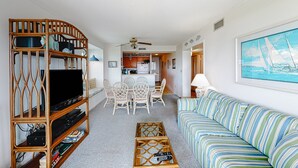 A living room with a TV on a wooden stand, a striped sofa, a dining area with chairs, and a kitchen in the background. - 2nd Floor in Resort w/Direct Beach Access + Free WiFi, Balcony, Shared Pool (Atlantic Beach)