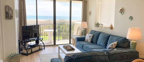 A living room with a sofa, TV, and a balcony with a view of the sea.