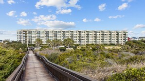 A wooden boardwalk leading to a modern multi-story building with balconies, surrounded by dense shrubbery. - Beachfront condo with amazing ocean views, pool, tennis courts, & gym (Atlantic Beach)
