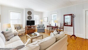 Uma sala de estar com sofá, poltronas, mesa de centro, televisão e piso de madeira.