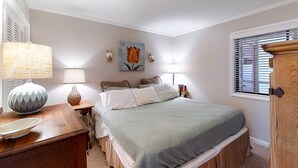 A bedroom with a bed, bedside lamps, a nightstand, a painting, and a window with blinds. - Lovely Condo in Direct Oceanfront Complex w/Shared Pool, Free WiFi, Water Views (Atlantic Beach)