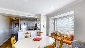 A dining area with a round table and chairs, a kitchen in the background, and a window with blinds. - Lovely Condo in Direct Oceanfront Complex w/Shared Pool, Free WiFi, Water Views (Atlantic Beach)