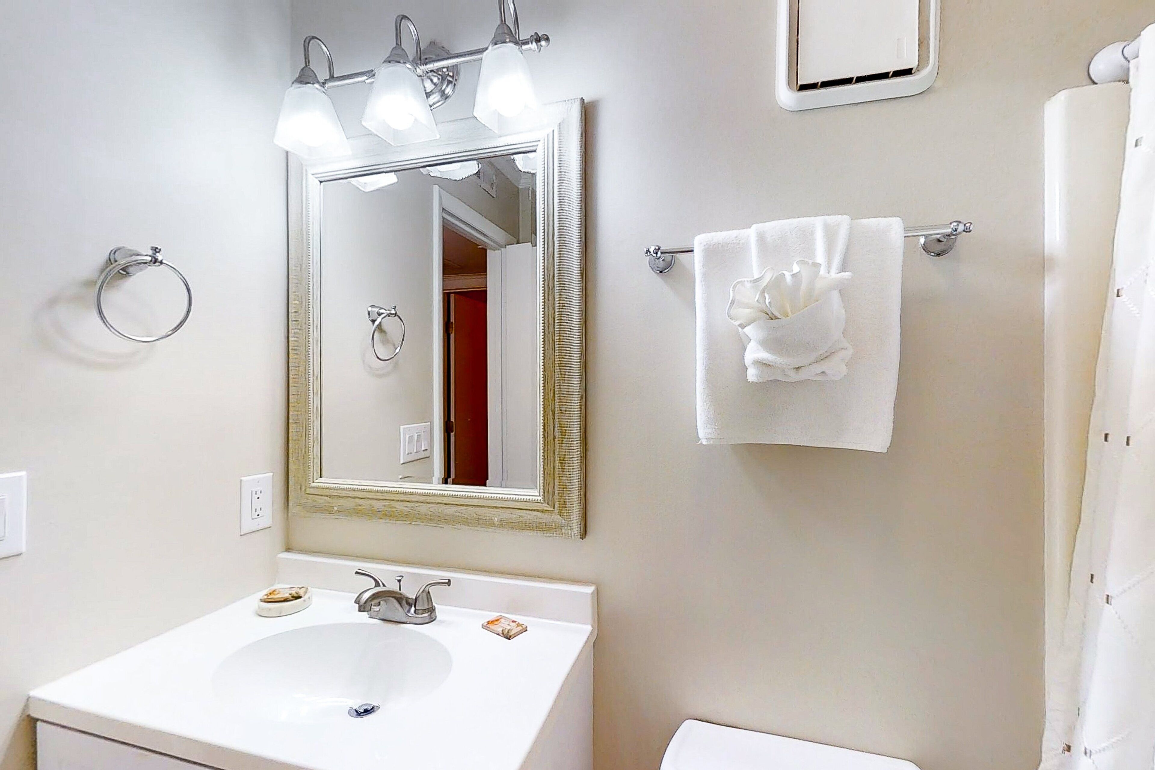 Une salle de bain avec un lavabo blanc, un miroir avec éclairage intégré, un porte-serviettes et un rideau de douche.