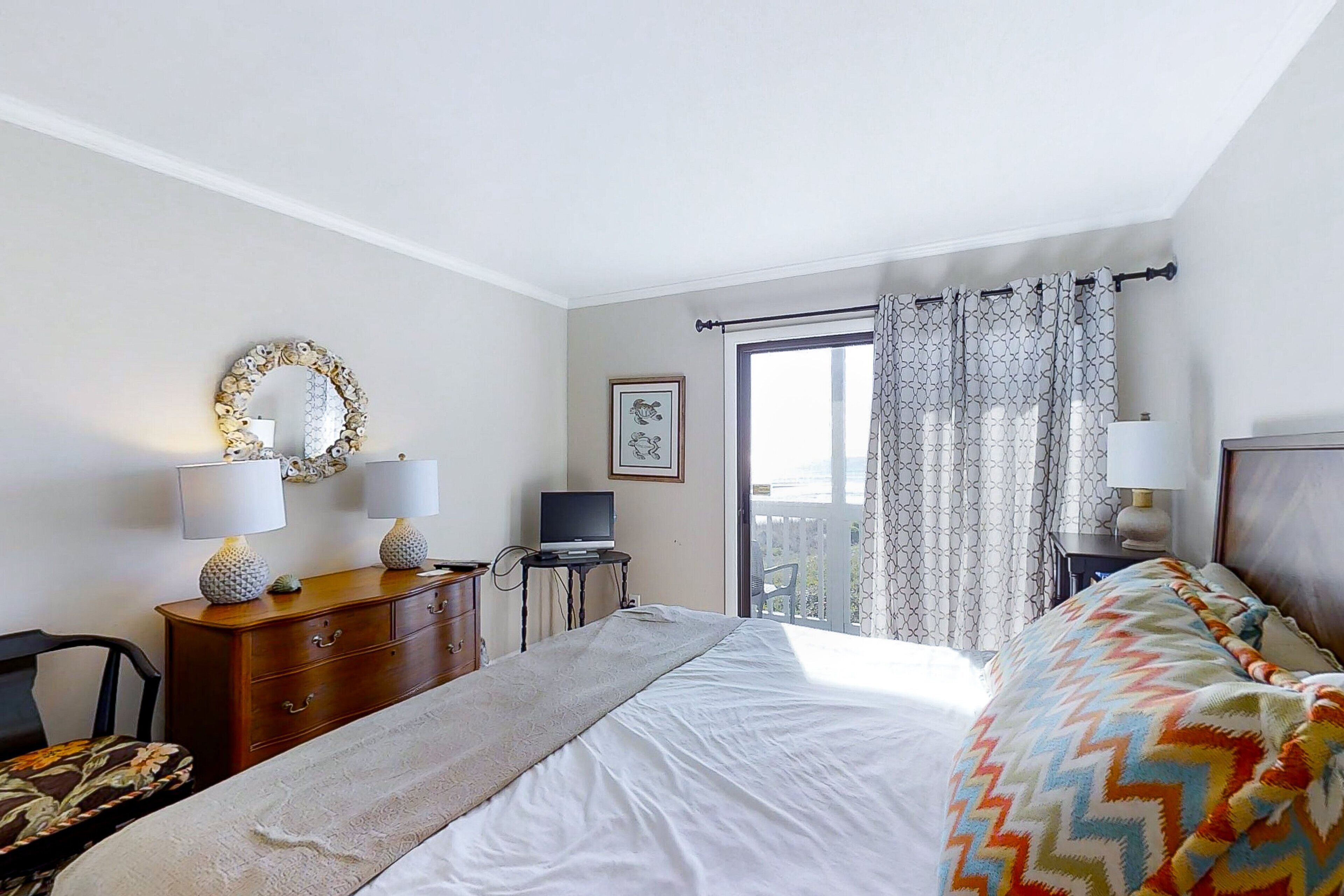 A hotel room with a bed, two bedside tables with lamps, a dresser, a television, and a balcony with curtains.