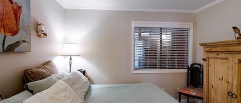 A bedroom with a bed, a nightstand, a lamp, a window with blinds, a chair, and a wooden cabinet.