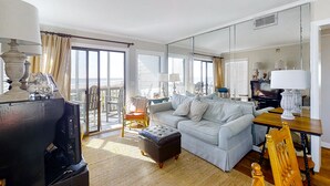 A living room with a sofa, ottoman, and a television. There is a dining table with a lamp, a chair, and a large mirror on the wall. - Lovely Condo in Direct Oceanfront Complex w/Shared Pool, Free WiFi, Water Views (Atlantic Beach)