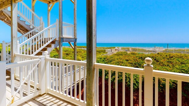 Une terrasse en bois, dotée de rampes blanches et d’un escalier menant à une zone abritée d’où l’on a vue sur la plage.