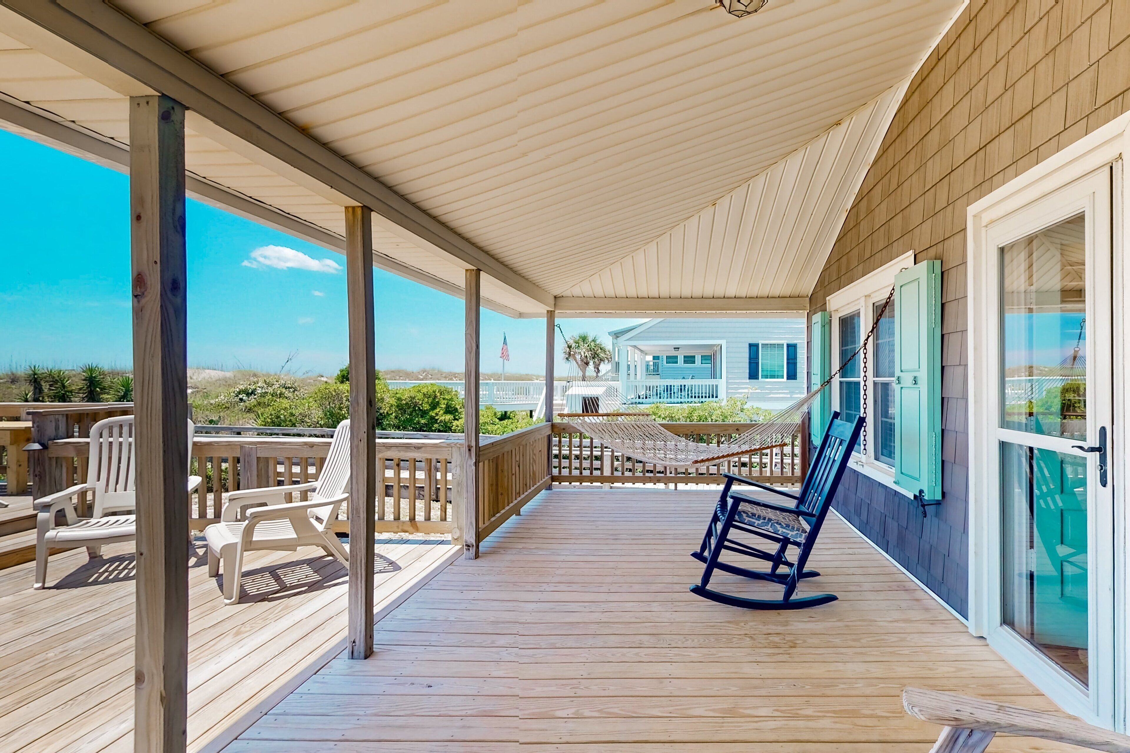 Eine Holzterrasse mit weißen Stühlen und einem Schaukelstuhl, mit Blick auf einen Strand und ein Haus mit blauen Fensterläden.
