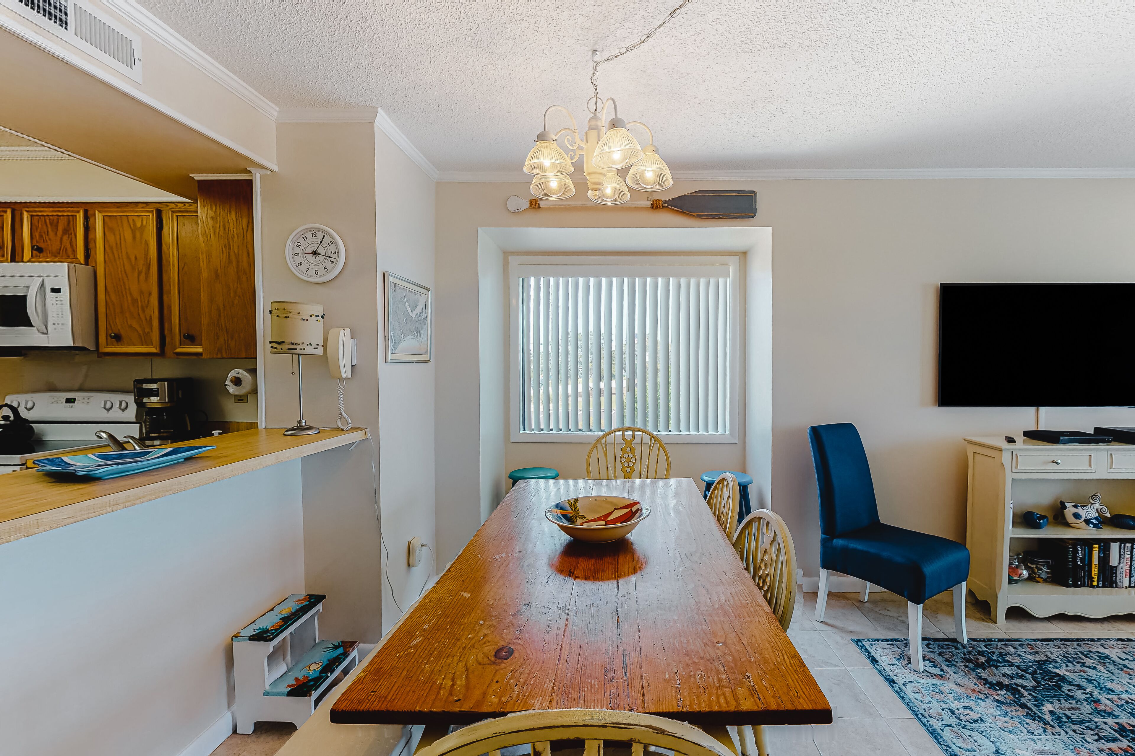 Un coin repas avec une table en bois, des chaises, un saladier de fruits, une horloge et un téléviseur en arrière-plan.