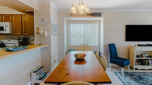 A dining area with a wooden table, chairs, a bowl of fruit, a clock, and a television in the background. - First-floor, oceanfront condo with great pool view, balconies, WiFi, shared pool (Atlantic Beach)