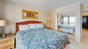 A bedroom with a large bed, a nightstand, a lamp, a wooden headboard, a painting, and a view through an open door. - First-floor, oceanfront condo with great pool view, balconies, WiFi, shared pool (Atlantic Beach)