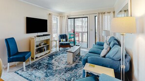 A living room with a blue sofa, a flat-screen TV, a lamp, and a bookshelf. - First-floor, oceanfront condo with great pool view, balconies, WiFi, shared pool (Atlantic Beach)