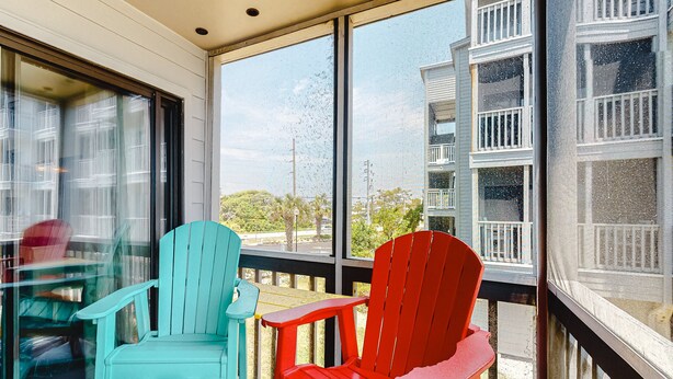 A balcony with two Adirondack chairs, one teal and one red, overlooking a residential area with houses and greenery. - First-floor, oceanfront condo with great pool view, balconies, WiFi, shared pool (Atlantic Beach)