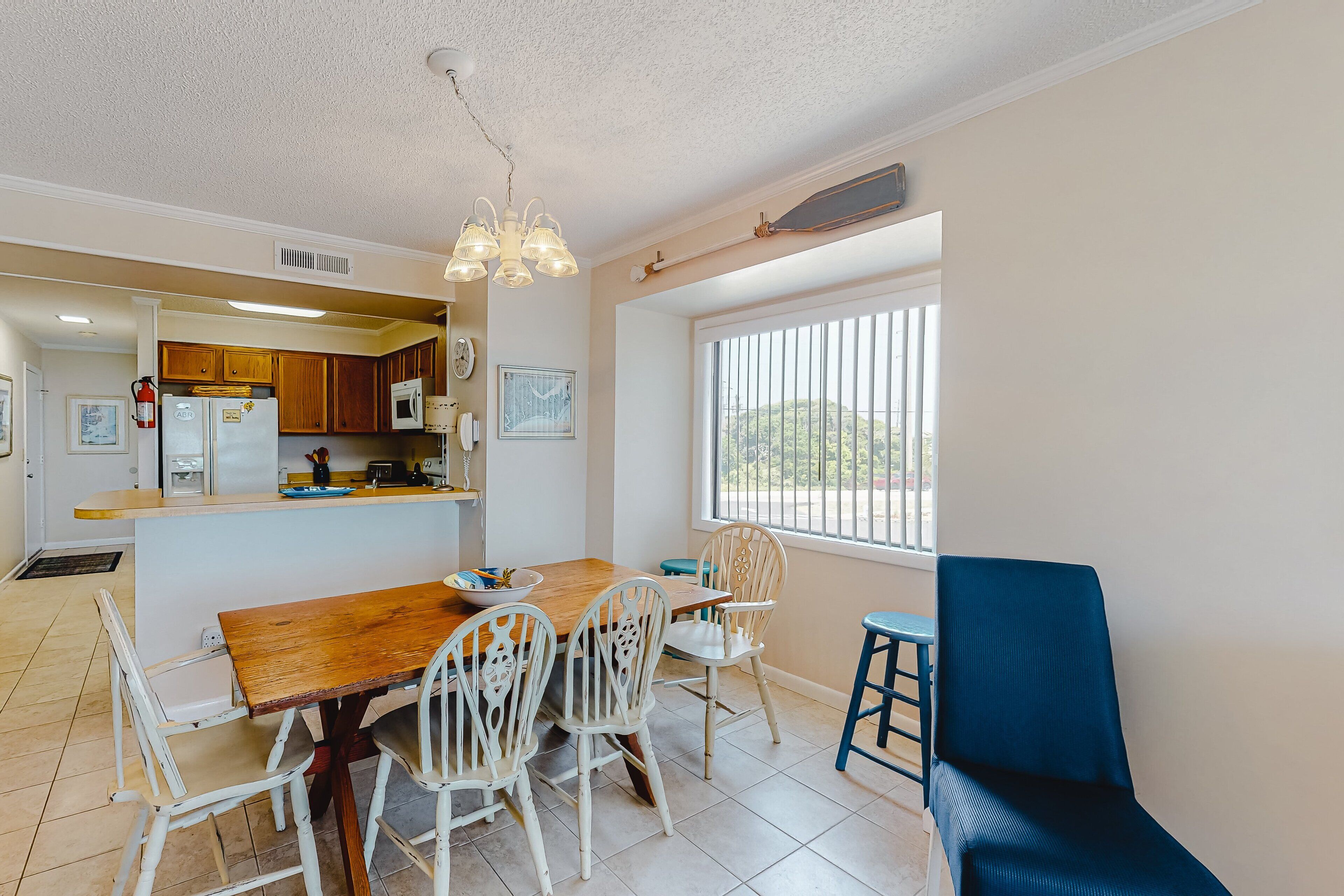 A kitchen with wooden cabinets, a dining table with white chairs, a blue stool, and a window with vertical blinds.