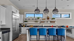 Een moderne keuken met witte kasten, een roestvrijstalen koelkast, een inbouwoven en een groot eiland met blauwe barkrukken. Boven het eiland hangen hanglampen en er is een raam met uitzicht op de lucht.