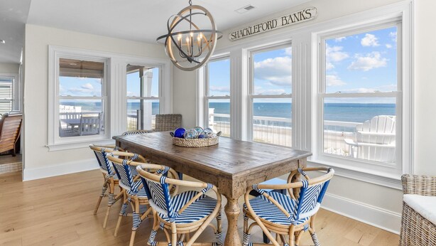 A dining area with a wooden table and wicker chairs, large windows with ocean views, and a chandelier.