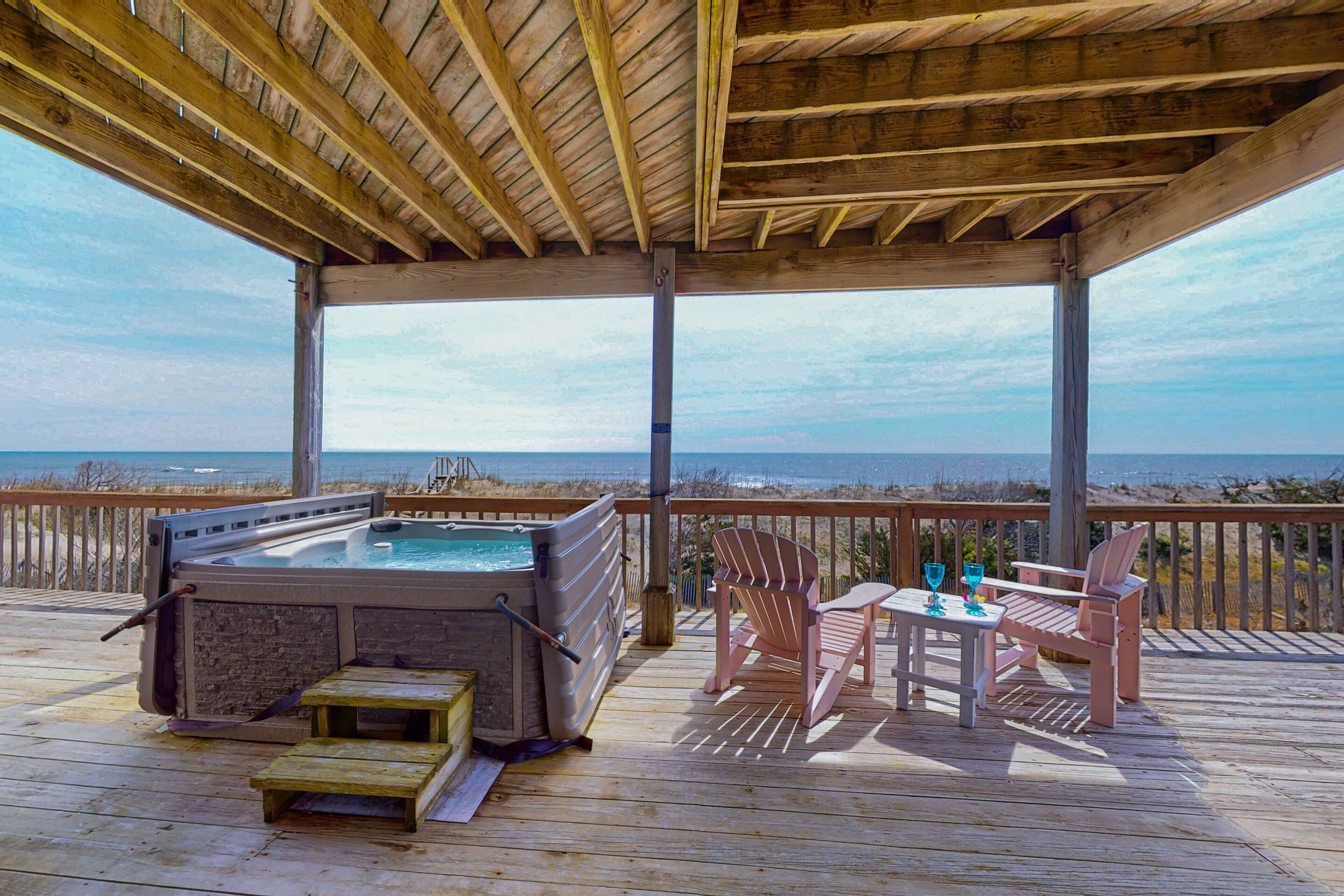 Een houten terras met een jacuzzi, stoelen en een tafel, met uitzicht op het strand.