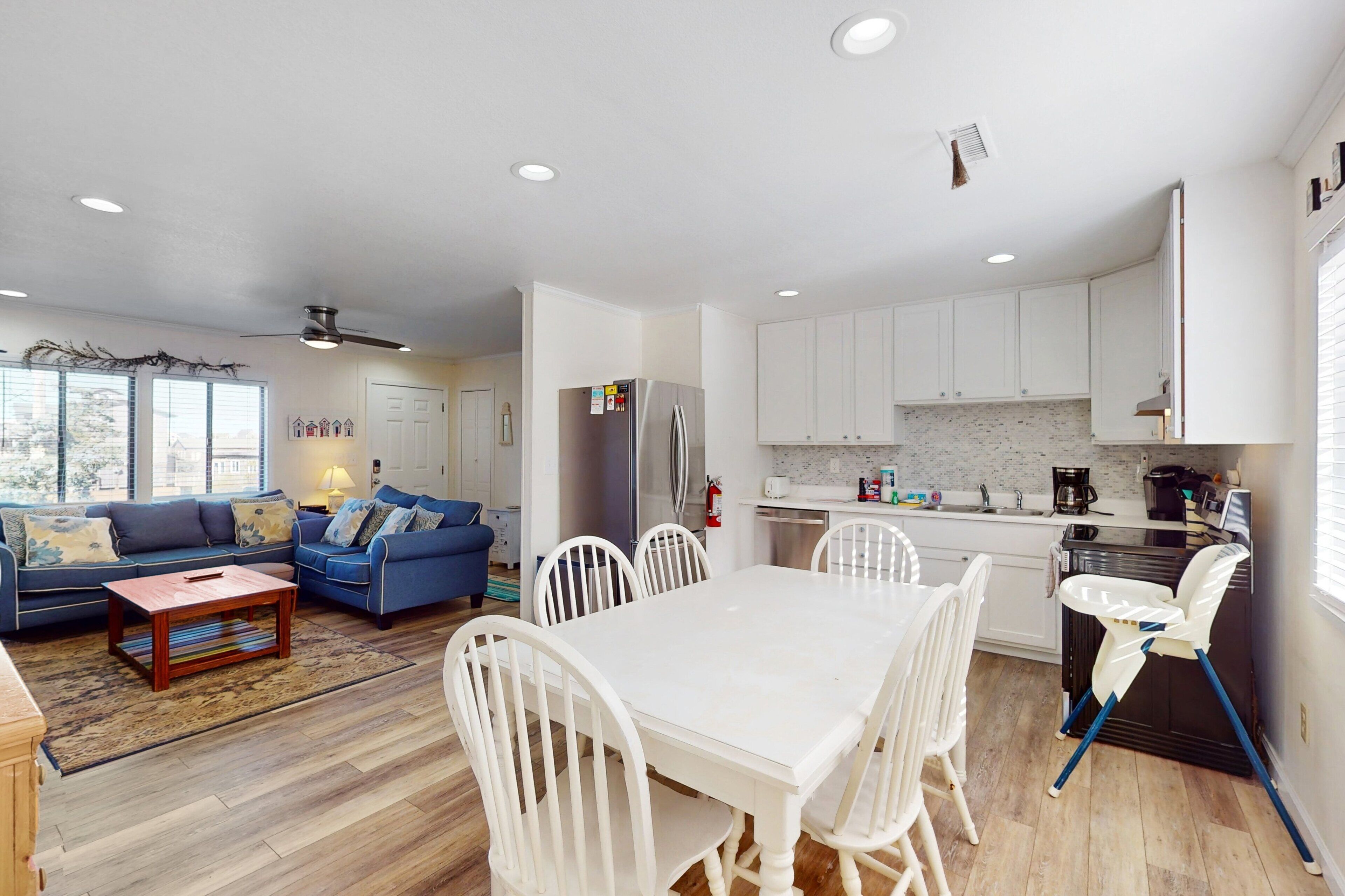 A modern living room with a blue sofa, a white dining table with chairs, and a kitchen area with white cabinets and a black countertop.