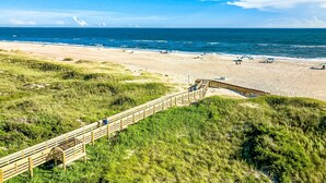 Una playa con un paseo marítimo de madera que conduce a la orilla, dunas cubiertas de hierba y el océano al fondo.