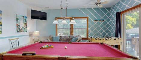 A pool table with a red felt surface, a ceiling fan, and a window with a view of trees.