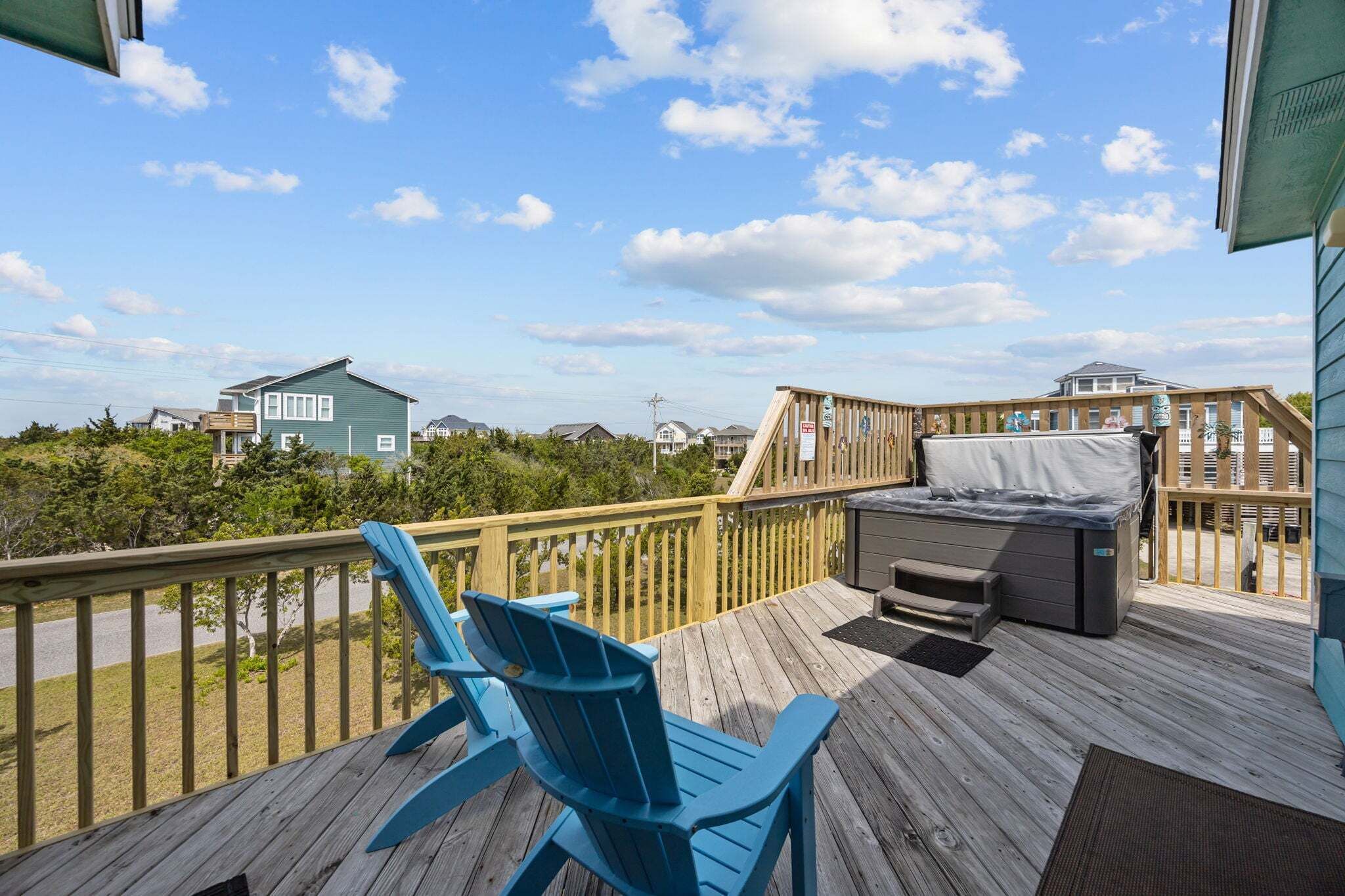 A wooden deck with a grill, a blue Adirondack chair, and a view of houses and trees.