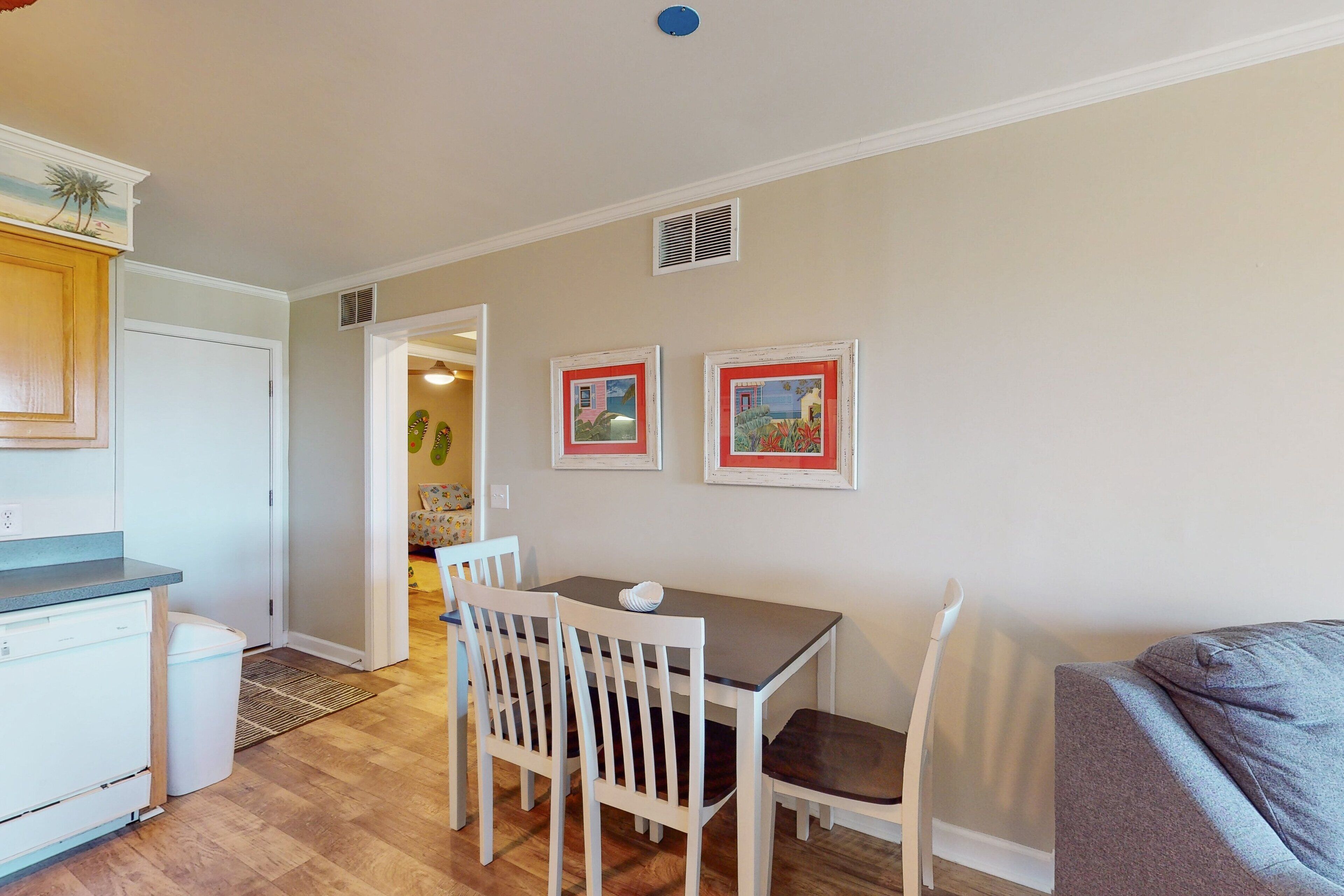 A compact kitchen with white cabinets, a dining table with four chairs, and two framed pictures on the wall.
