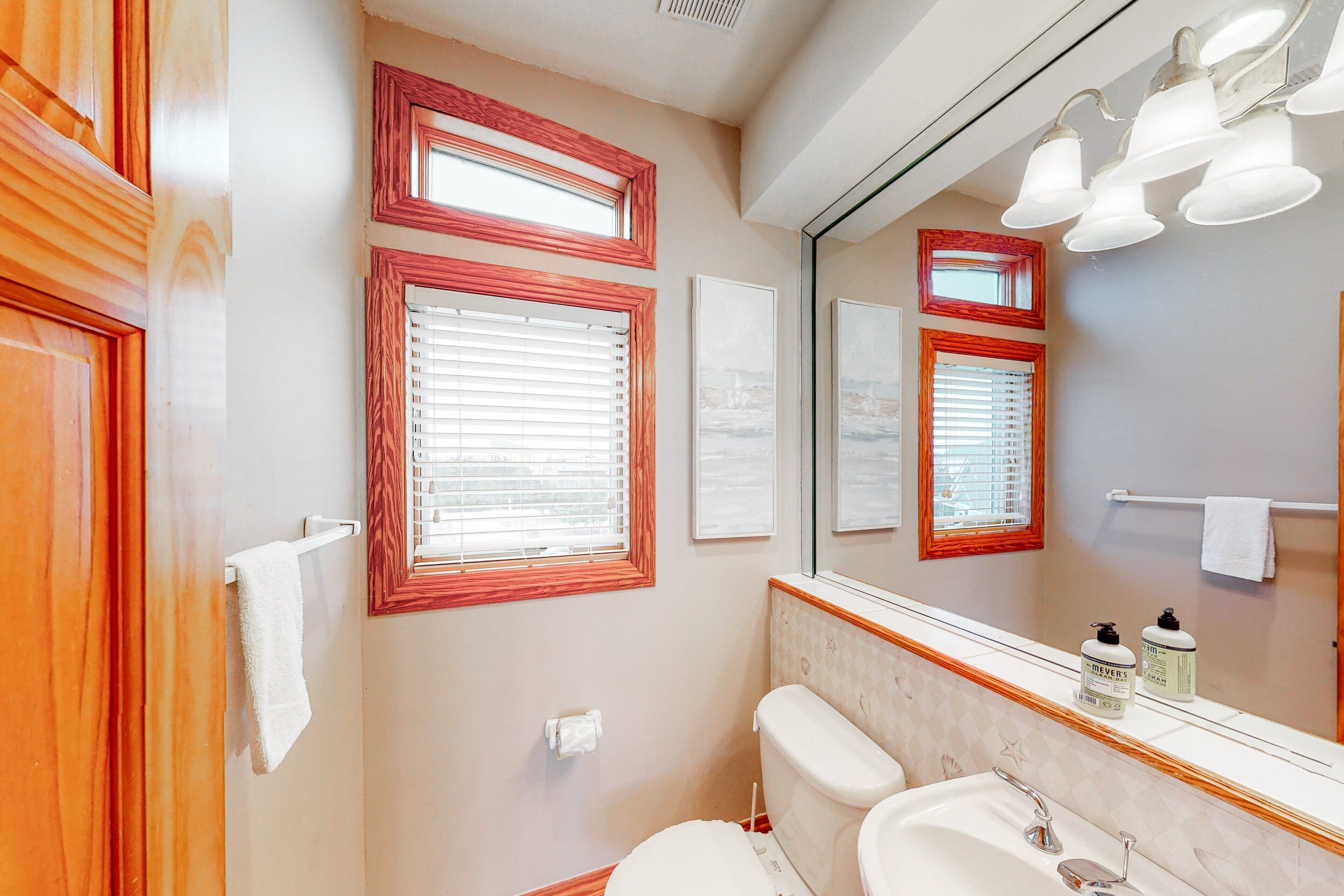 A bathroom with two sinks, a toilet, and a large mirror. There are three windows with wooden frames, and a towel rack is visible.