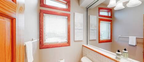 A bathroom with two sinks, a toilet, and a large mirror. There are three windows with wooden frames, and a towel rack is visible.