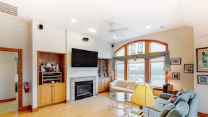 A spacious living room with a fireplace, a ceiling fan, and a large window with curtains.