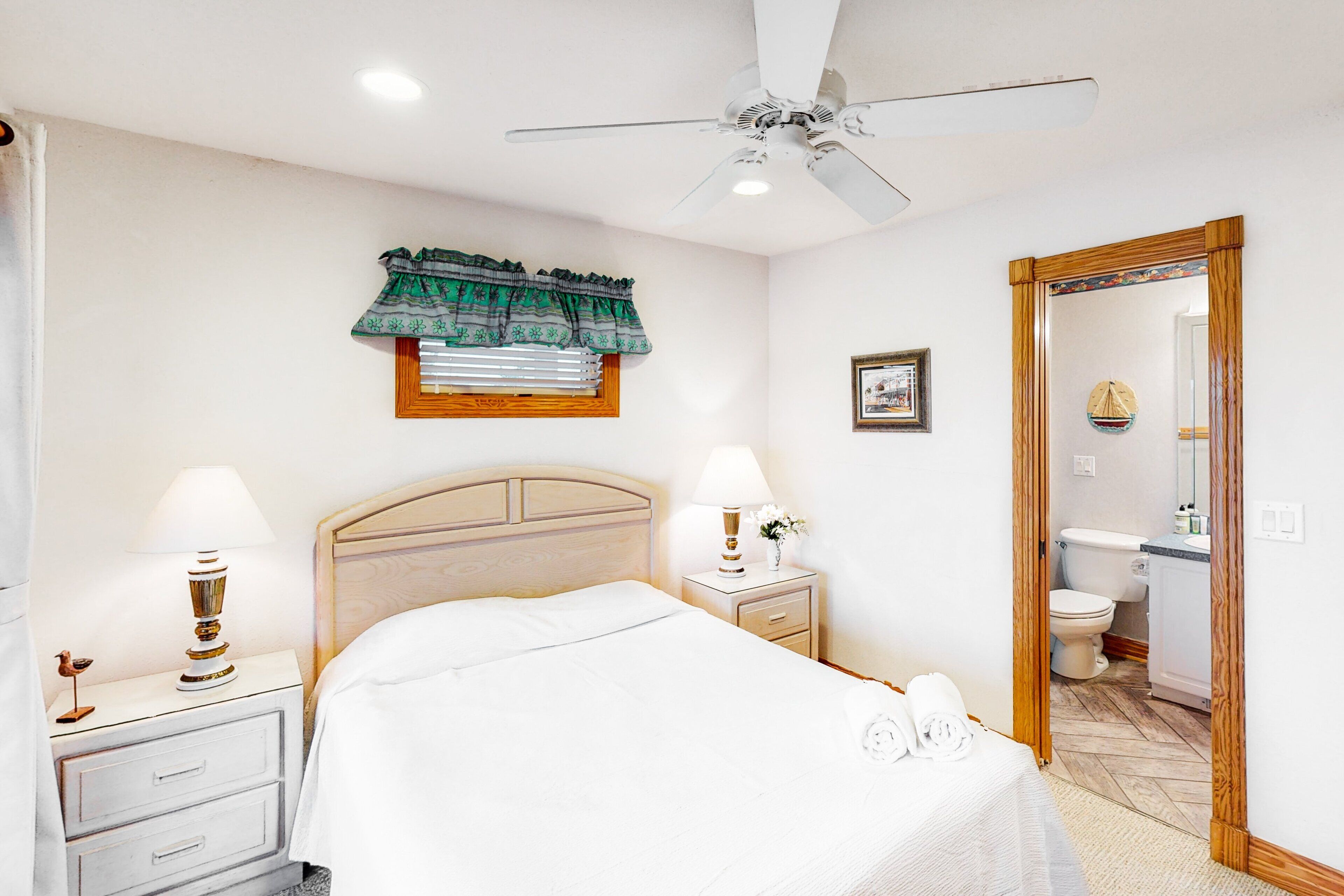 A bedroom with a bed, two bedside tables with lamps, a ceiling fan, a window with a curtain, and a door leading to a bathroom.