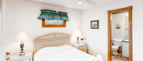 A bedroom with a bed, two bedside tables with lamps, a ceiling fan, a window with a curtain, and a door leading to a bathroom.