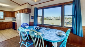 Un coin repas avec une table dressée pour quatre personnes, des meubles en bois, un réfrigérateur et une vue sur les maisons à travers de grandes fenêtres.