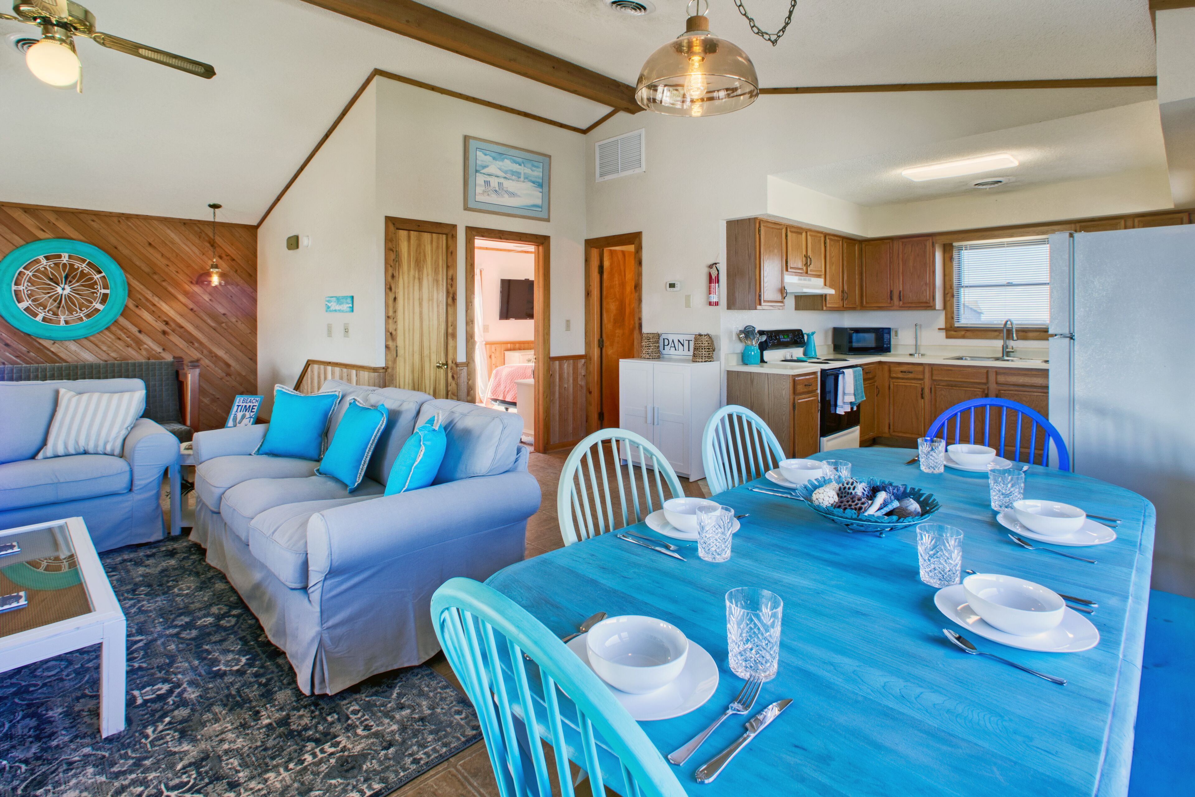 A modern kitchen with wooden cabinets, a microwave, and a refrigerator. There is a dining table set with white plates, bowls, and cutlery. The room has a blue tablecloth and light blue chairs.